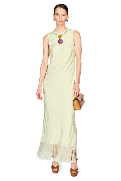 A model wearing a sleeveless, pale yellow-green maxi dress with a sheer tulle hem, accessorized with a colorful pendant necklace and a brown handbag, set against a plain white background.