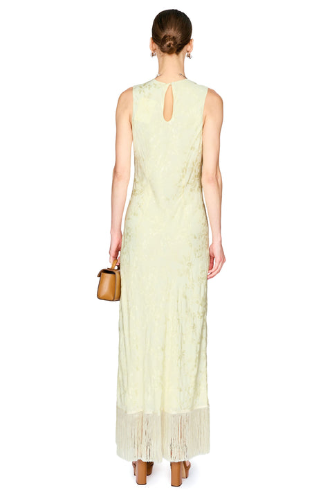 A woman viewed from the back wearing a sleeveless pale yellow-green maxi dress with a small keyhole cutout at the back and fringe trim at the hem. She is standing against a plain white background and holding a small tan handbag.