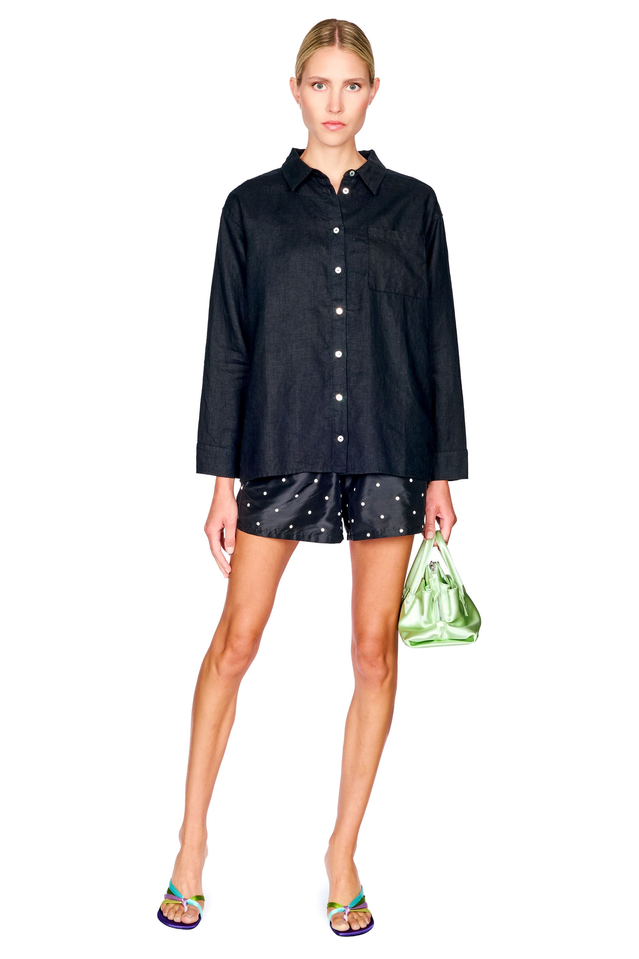 A model stands against a white background wearing a black long-sleeve button-up shirt, black shorts with white polka dots, and holding a green patterned handbag.