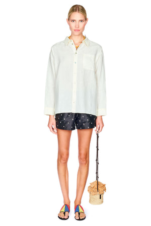 A model stands against a white background wearing a light-colored, oversized long-sleeve button-down shirt, dark patterned shorts, and colorful sandals, while holding a small woven bucket bag.