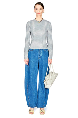 A model stands against a white background wearing a gray and white striped long-sleeve top, wide-leg blue jeans, beige sandals, and holding a light beige handbag.