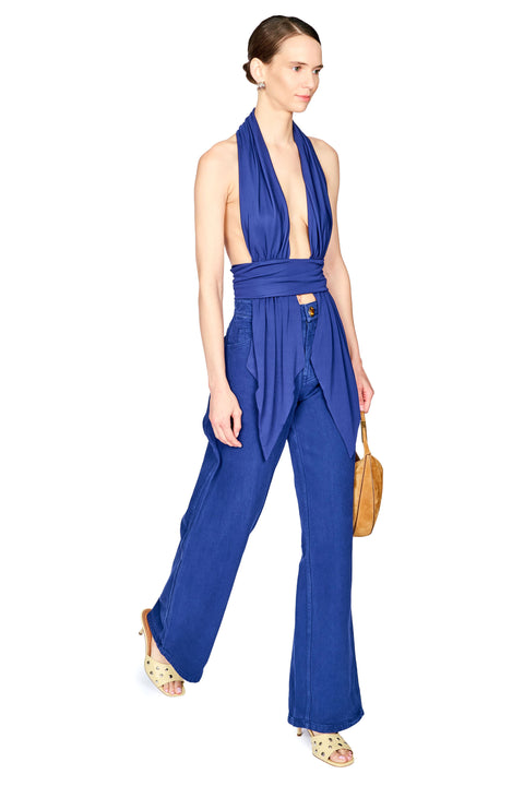 A model wearing a royal blue halter-neck top with wide-leg pants, holding a tan leather handbag against a plain white background.