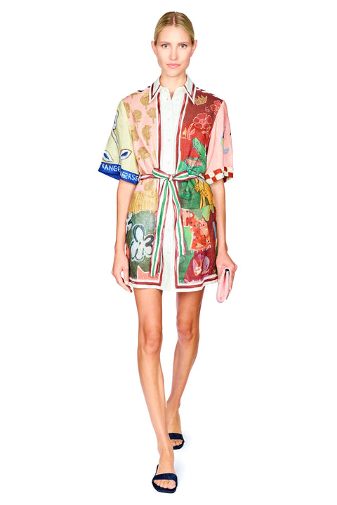 A model walks against a white background wearing a short-sleeved, collared mini dress with a colorful patchwork print, featuring green, red, pink, and beige tones, with a white shirt and navy blue sandals.