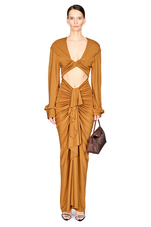 A woman on a white background wearing a cognac brown long dress with long sleeves, drapery from the waist down, and a cutout under the chest
