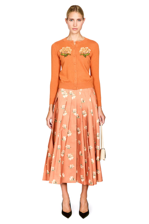 A model stands against a white background wearing a two-piece outfit consisting of a burnt orange long-sleeve button-up cardigan with floral embroidery on the chest and a matching peach pleated midi skirt with a white floral print. She is wearing black pointed-toe heels and holding a small beige handbag.