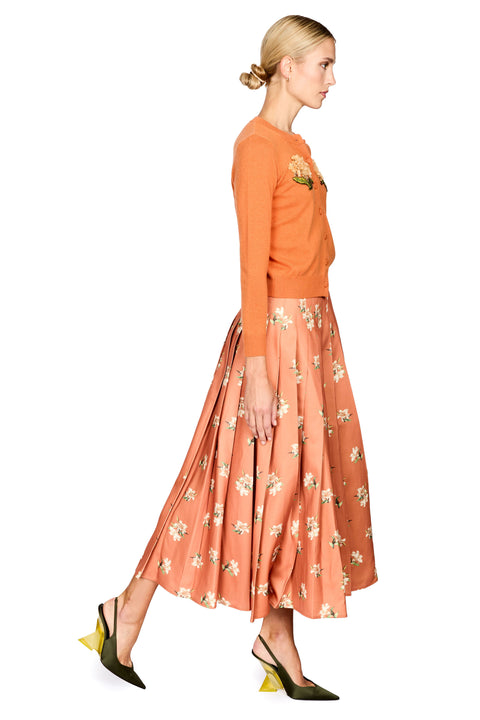 A woman in profile wearing a rust-orange long-sleeve knit top and a pleated peach skirt with a white floral print, paired with olive green pointed-toe heels, against a plain white background.