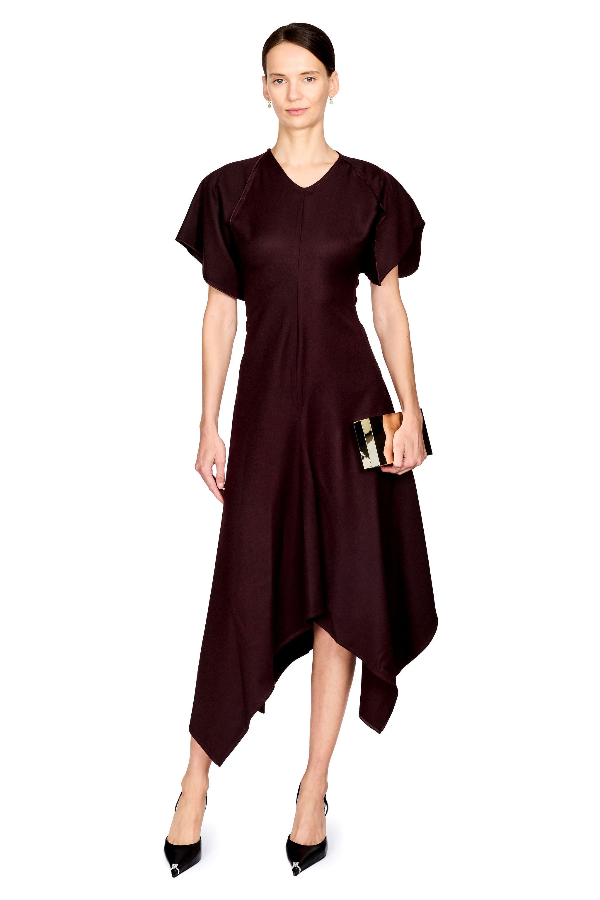 A woman modeling a dark burgundy midi dress with short puff sleeves, a V-neckline, and an asymmetrical draped hem. She is holding a small black clutch and wearing black pointed-toe heels against a plain white background.