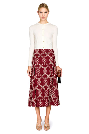 A woman modeling a white long-sleeve knit top and a burgundy midi skirt with a cream geometric trellis pattern, holding a small black clutch against a plain white background.