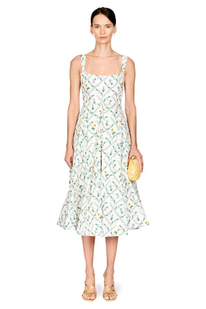 A model stands against a white studio background wearing a sleeveless midi dress with a white base and a green and yellow botanical pattern. She is holding a small yellow clutch and wearing gold sandals.