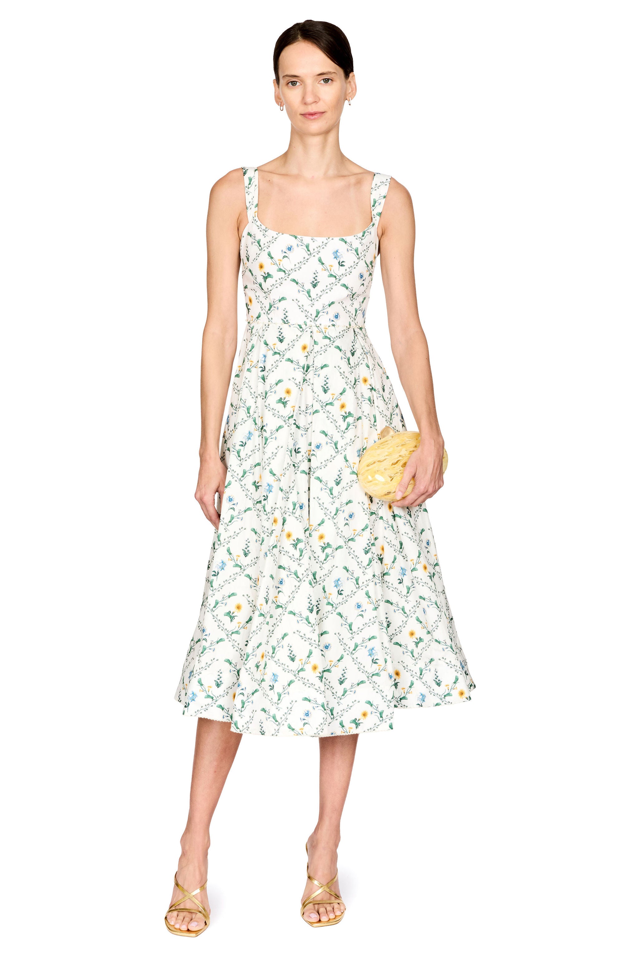 A woman modeling a sleeveless midi dress with a white base and a green, yellow, and orange floral print, holding a small yellow clutch against a plain white background.