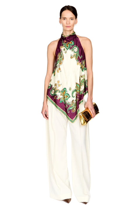 A model stands against a white studio background wearing a sleeveless high-neck top made from a square scarf with a green, gold, and burgundy paisley print over an off-white base, paired with wide-leg off-white pants and holding a gold clutch.