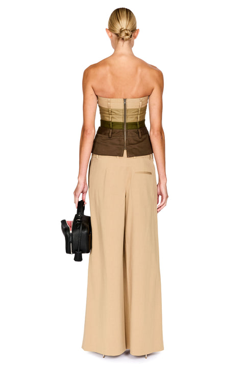 A model shown from the back wearing a strapless two-tone dress with a structured corset-style bodice in olive green and dark brown, paired with wide-leg beige trousers. She is holding a small black handbag against a plain white background.