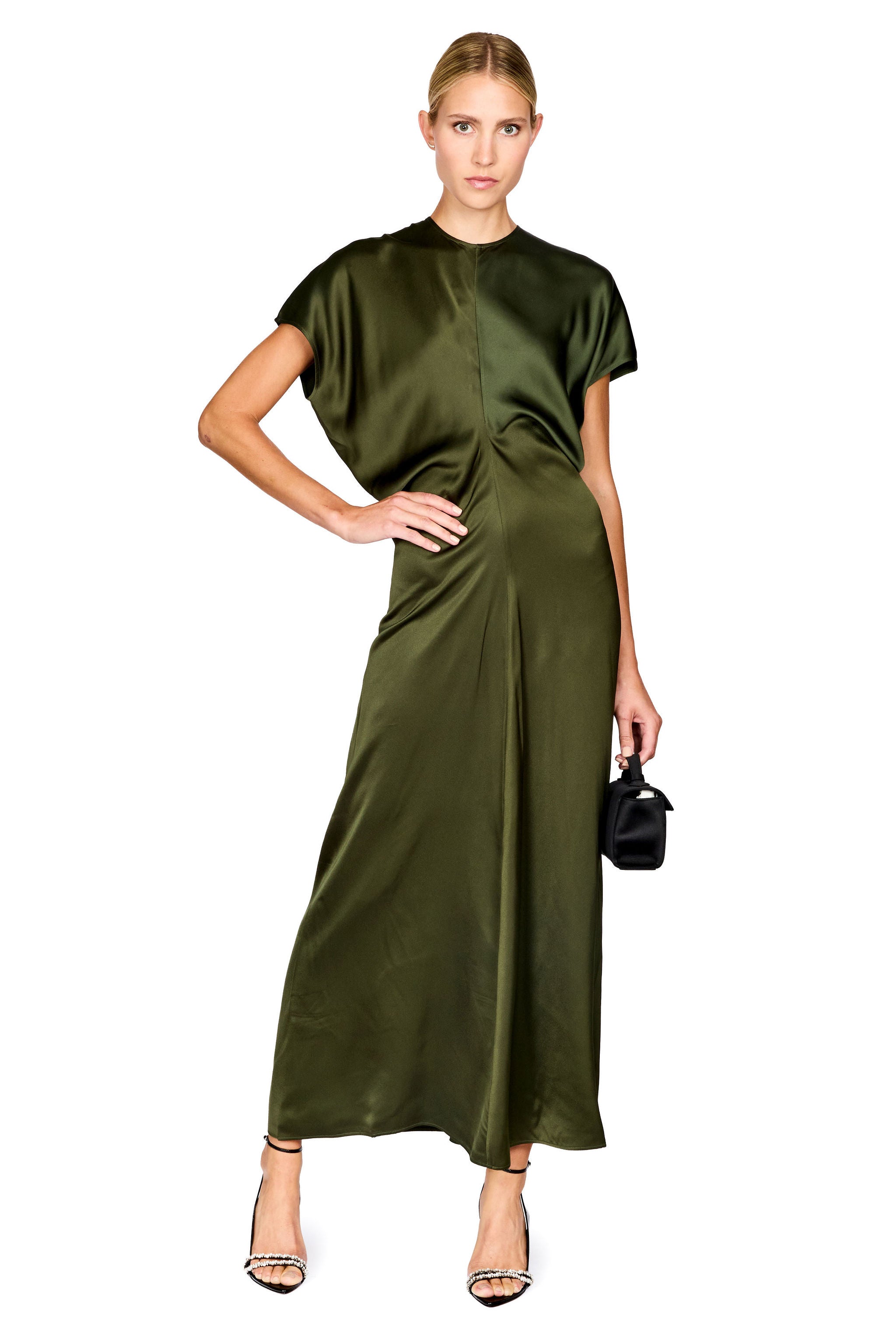 A woman modeling a short-sleeved olive green satin maxi dress with a draped, asymmetrical front design, paired with black high-heeled sandals and a small black handbag, set against a plain white background.