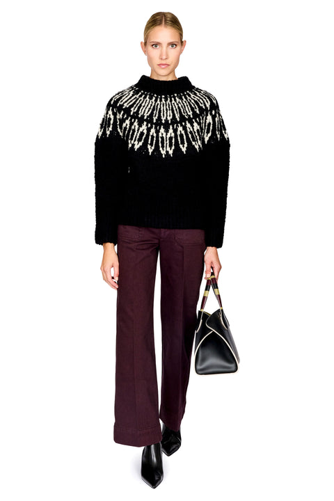 A model stands against a white background wearing a black knit sweater with a white geometric pattern across the chest, dark plum wide-leg trousers, black pointed shoes, and holding a black handbag.