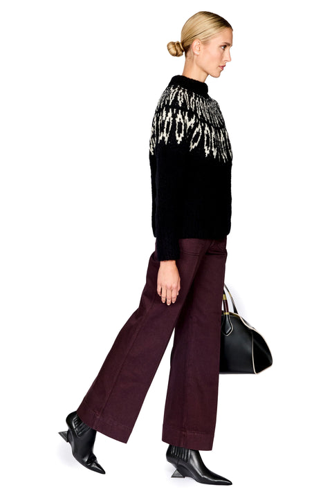 A woman modeling a black knit sweater with a white geometric pattern across the shoulders, paired with wide-leg burgundy trousers and black pointed-toe ankle boots, holding a black handbag against a plain white background.