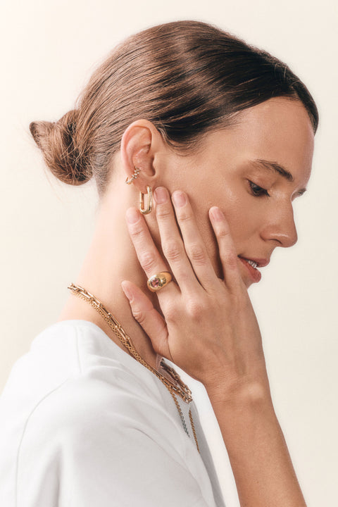 A woman in profile wearing gold jewelry, including a gold hoop earring, a gold ring, and a gold chain necklace, against a clean off-white background.