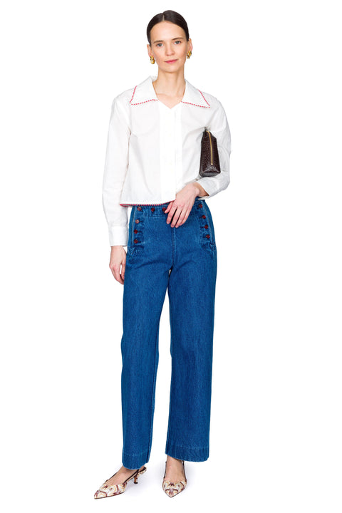 Woman wearing a white blouse with red accents and blue jeans with red buttons on a white background with one hand in pocket.