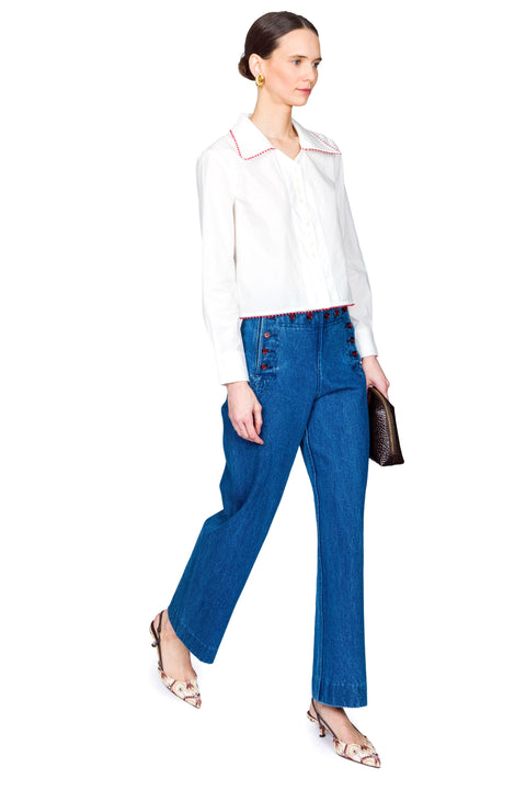 Woman wearing a white blouse with red accents and blue jeans with red buttons on a white background from a side view.