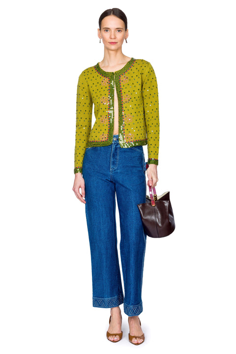 Woman wearing a green embroidered sweater and blue jeans with a brown handbag on a white background with hip to one side.