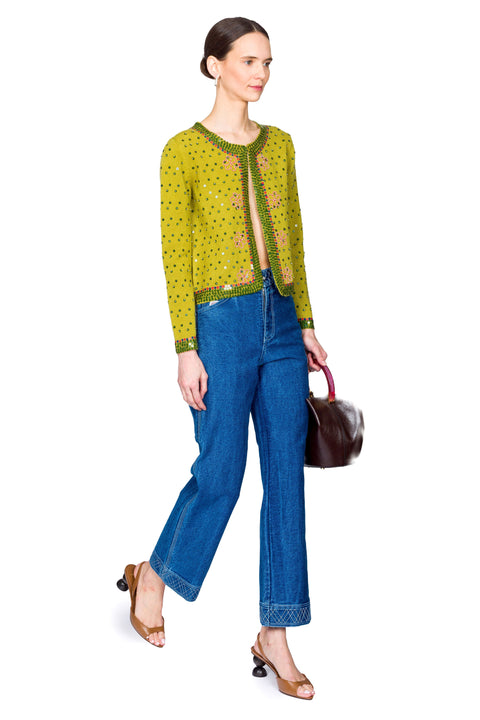 Woman wearing a green embroidered sweater and blue jeans with a brown handbag on a white background with one leg in front.