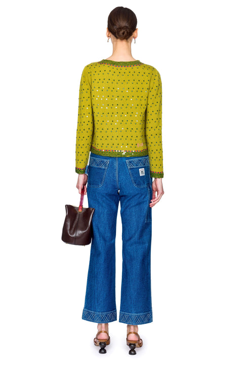 Woman wearing a green embroidered sweater and blue jeans with a brown handbag on a white background from the back.
