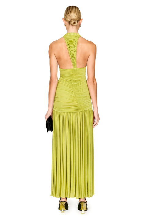 A woman viewed from the back wearing a sleeveless halter-neck maxi dress in a bright chartreuse/lime green fabric with ruching at the bodice and a long pleated skirt. She is standing against a plain white studio background and holding a small black clutch.