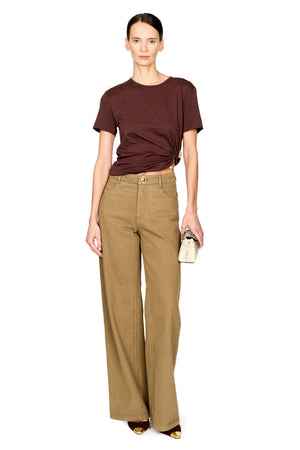 A model stands against a white background wearing a dark brown short-sleeve t-shirt with a twisted hem detail, high-waisted wide-leg tan trousers, and holding a small textured clutch.