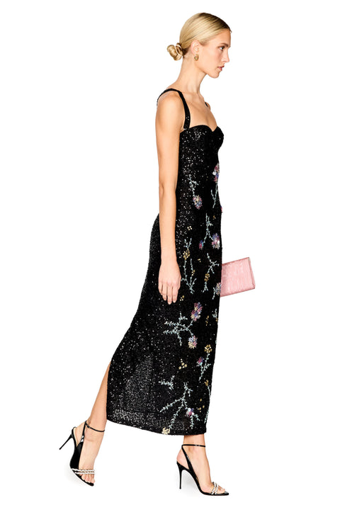 A woman modeling a black spaghetti-strap midi dress covered in sequins and floral embroidery, paired with black high-heeled sandals and a small pink clutch, shown against a plain white background.