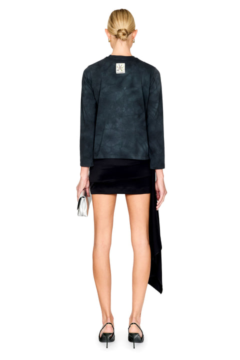 A model shown from the back wearing a dark charcoal tie-dye long-sleeve top and a black velvet mini skirt with an asymmetrical draped hem, paired with black heels and holding a silver clutch against a white background.