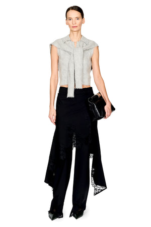 A model stands against a white studio background wearing a light gray sleeveless fuzzy wrap top with a tie detail, black wide-leg trousers with lace trim, and holding a small black clutch.