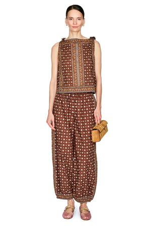 A model stands against a white studio background wearing a brown sleeveless top and matching wide-leg pants with a dainty pattern and gold trim, holding a small tan clutch.