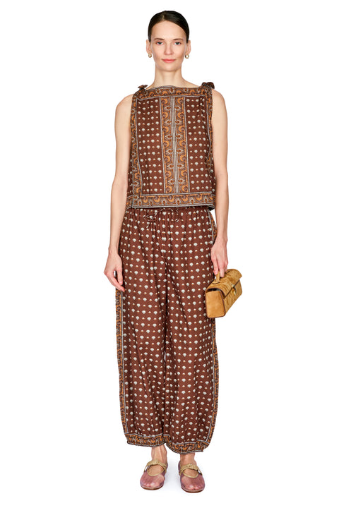 A model stands against a white studio background wearing a brown sleeveless top and matching wide-leg pants with a dainty pattern and gold trim, holding a small tan clutch.
