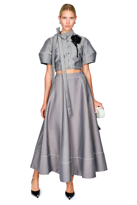 A model wearing a gray two-piece outfit consisting of a short-sleeve cropped top with a tie-neck detail and a matching flared midi skirt, styled with black pointed heels and a small white clutch against a white background.