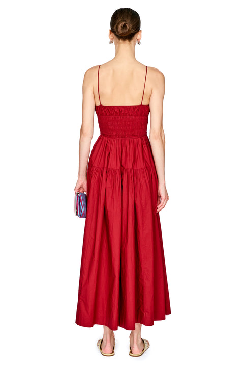 A woman viewed from the back wearing a sleeveless red maxi dress with thin straps, a smocked bodice, and a tiered skirt. She is standing against a plain white background and holding a small striped clutch.