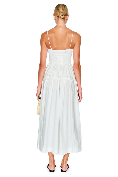 A woman viewed from the back wearing a white spaghetti-strap midi dress with a smocked bodice and a flowing skirt, paired with black slide sandals and holding a small woven clutch against a plain white background.