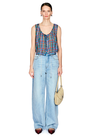 A model stands against a white background wearing a colorful plaid sleeveless top, light-wash wide-leg denim jeans, and holding a beige shoulder bag.