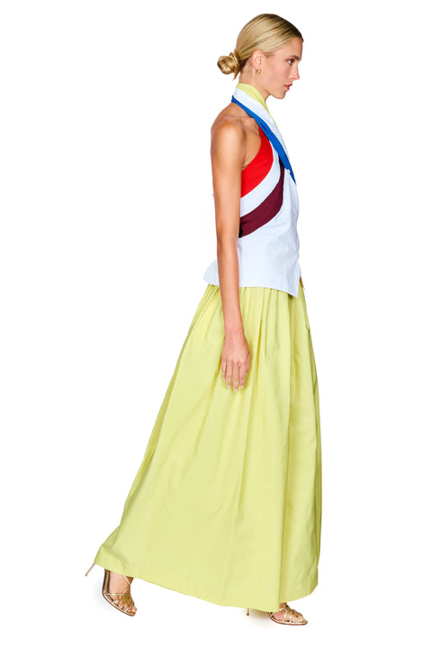 A model wearing a sleeveless halter top with diagonal red, yellow, blue, and white stripes, paired with a flowing pale yellow skirt, turned to the side