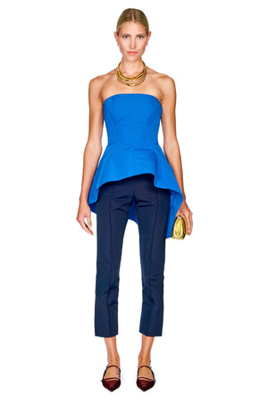 A model wearing a bright blue strapless peplum top and navy blue cropped trousers, accessorized with a gold necklace and holding a gold clutch, set against a plain white background.