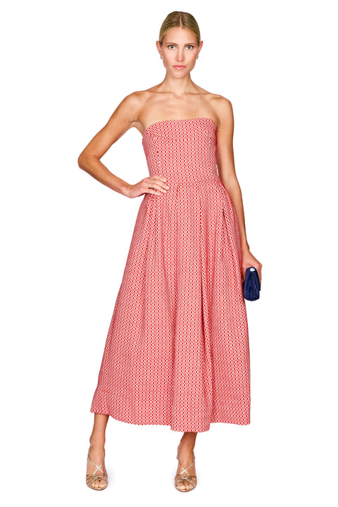 A woman modeling a strapless midi dress with a pink and white geometric pattern, holding a small navy blue clutch against a solid white background.