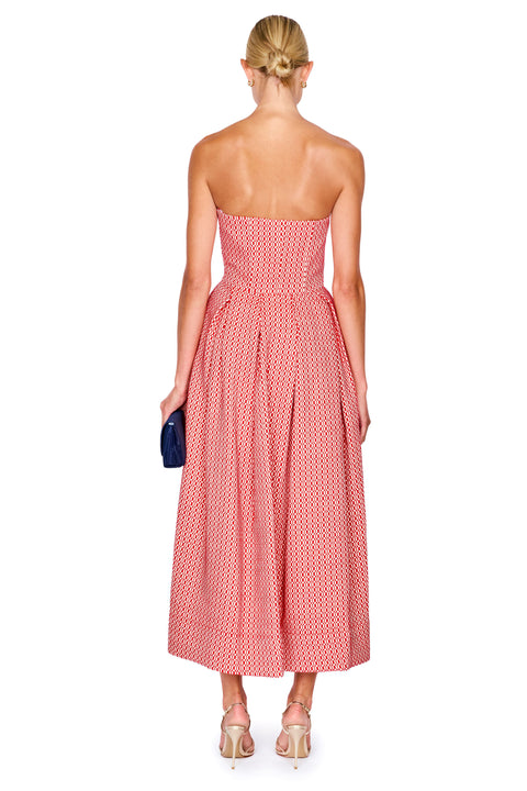 A woman viewed from the back wearing a strapless midi dress with a red and white geometric pattern, holding a small navy blue clutch.