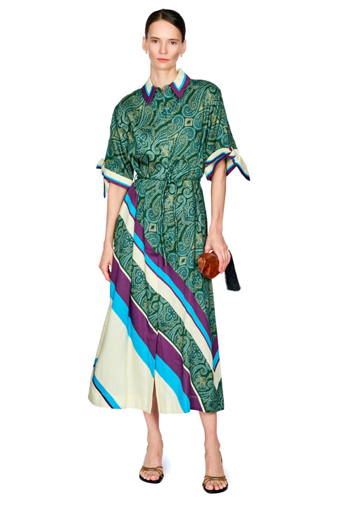 A model wearing a green paisley midi dress with diagonal stripes in blue, purple, and cream, styled with a small brown clutch and black sandals against a white background.