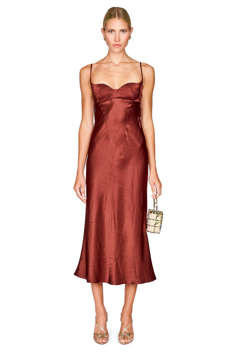 A woman modeling a satin slip midi dress in a rust brown color, styled with a small flower-patterned handbag and metallic heels, against a plain white background.