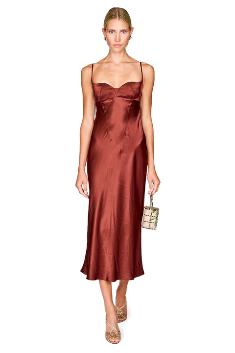 A woman modeling a satin slip dress in a rust-brown color, styled with a small snakeskin-patterned handbag and clear high heels, against a plain white background.