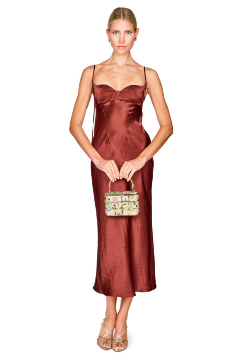 A woman modeling a satin slip dress in a deep rust-brown color, styled with a small patterned handbag and metallic heels against a plain white background.