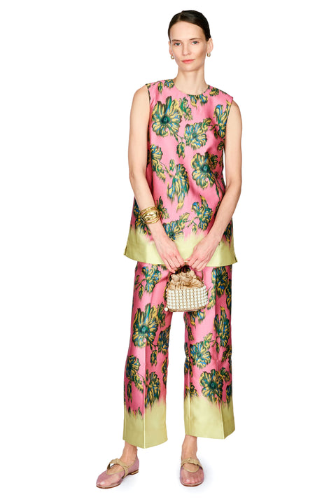 A model stands against a white studio background wearing a sleeveless pink top and pants set with a green and blue floral print and a light yellow-green gradient hem. She is holding a small woven handbag and wearing gold bracelets and pink shoes.