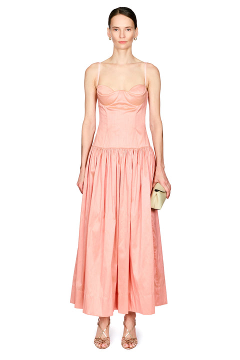 A model wearing a light pink spaghetti-strap maxi dress with a structured bodice and a flowing skirt, holding a small beige clutch against a plain white background.