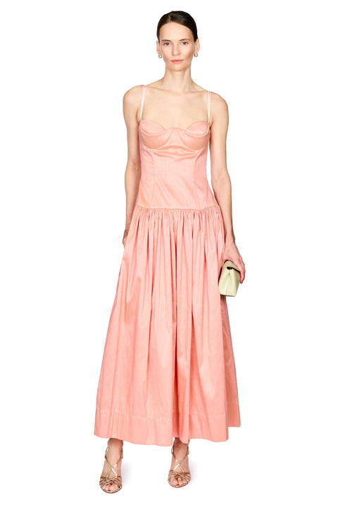A model wearing a light pink structured dress with spaghetti straps and a gathered skirt, holding a small beige clutch against a plain white background.