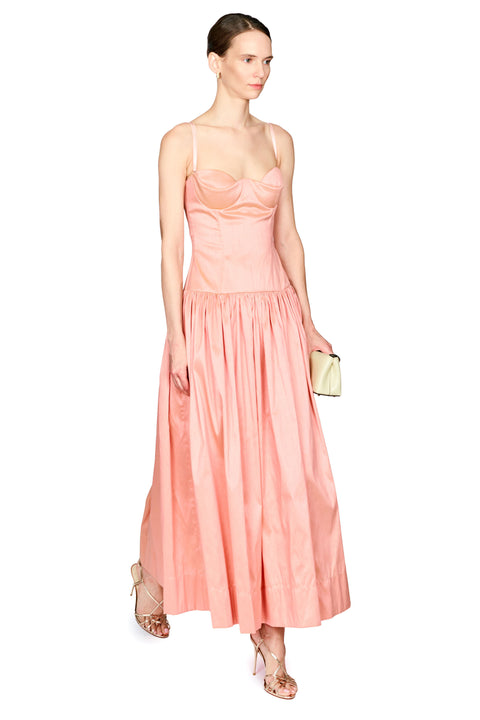 A woman modeling a peach-colored maxi dress with spaghetti straps, a structured sweetheart bodice, and a full gathered skirt, holding a small cream clutch.
