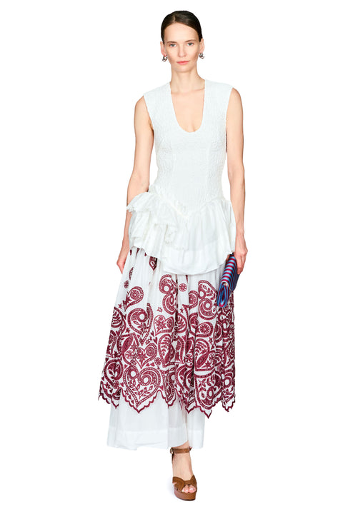 A model wearing a sleeveless white top with a ruffled waist and a long white skirt featuring a burgundy paisley pattern and scalloped hem, styled with brown sandals against a plain white background.
