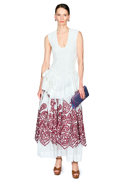 A model wearing a sleeveless white ruffled top and a long white skirt with a burgundy paisley print, holding a blue striped clutch against a plain white background.